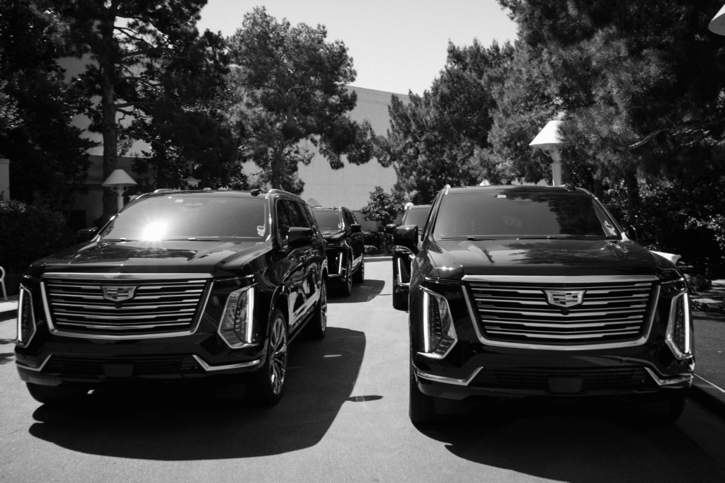 Black-and-white photo of two black Cadillac Escalades parked front-facing in a tree-lined driveway, with additional fleet vehicles visible behind them.