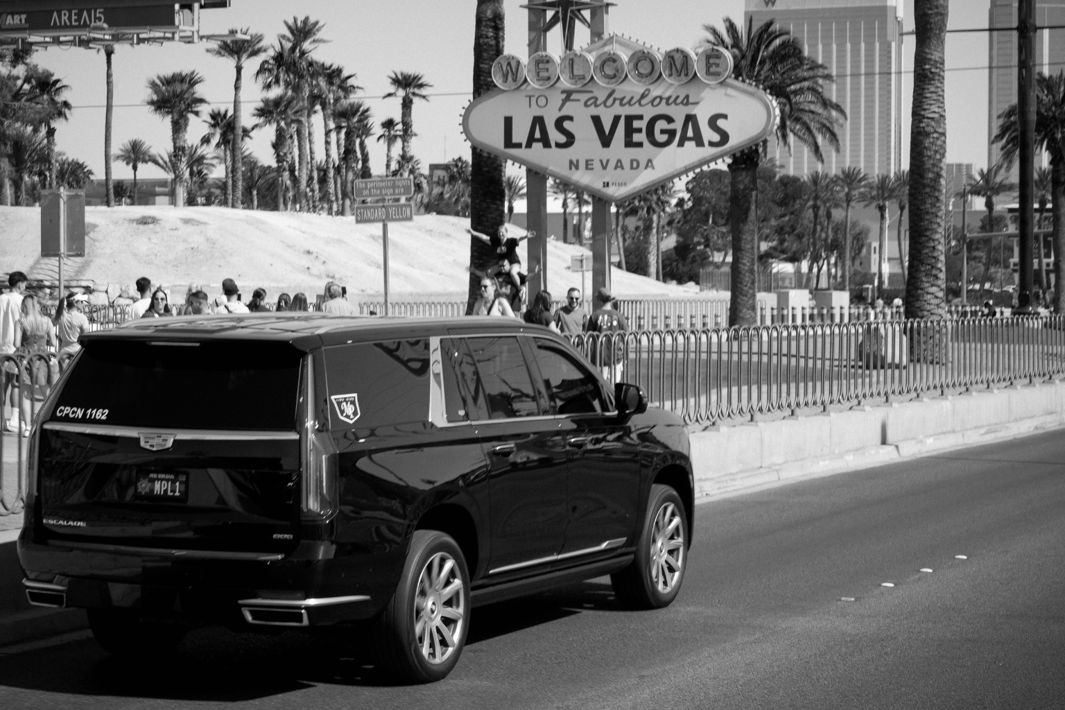 Black-and-white photo of a black Cadillac Escalade with MP Limousine branding and an MPL1 license plate driving past the iconic "Welcome to Fabulous Las Vegas Nevada" sign, with tourists gathered nearby.