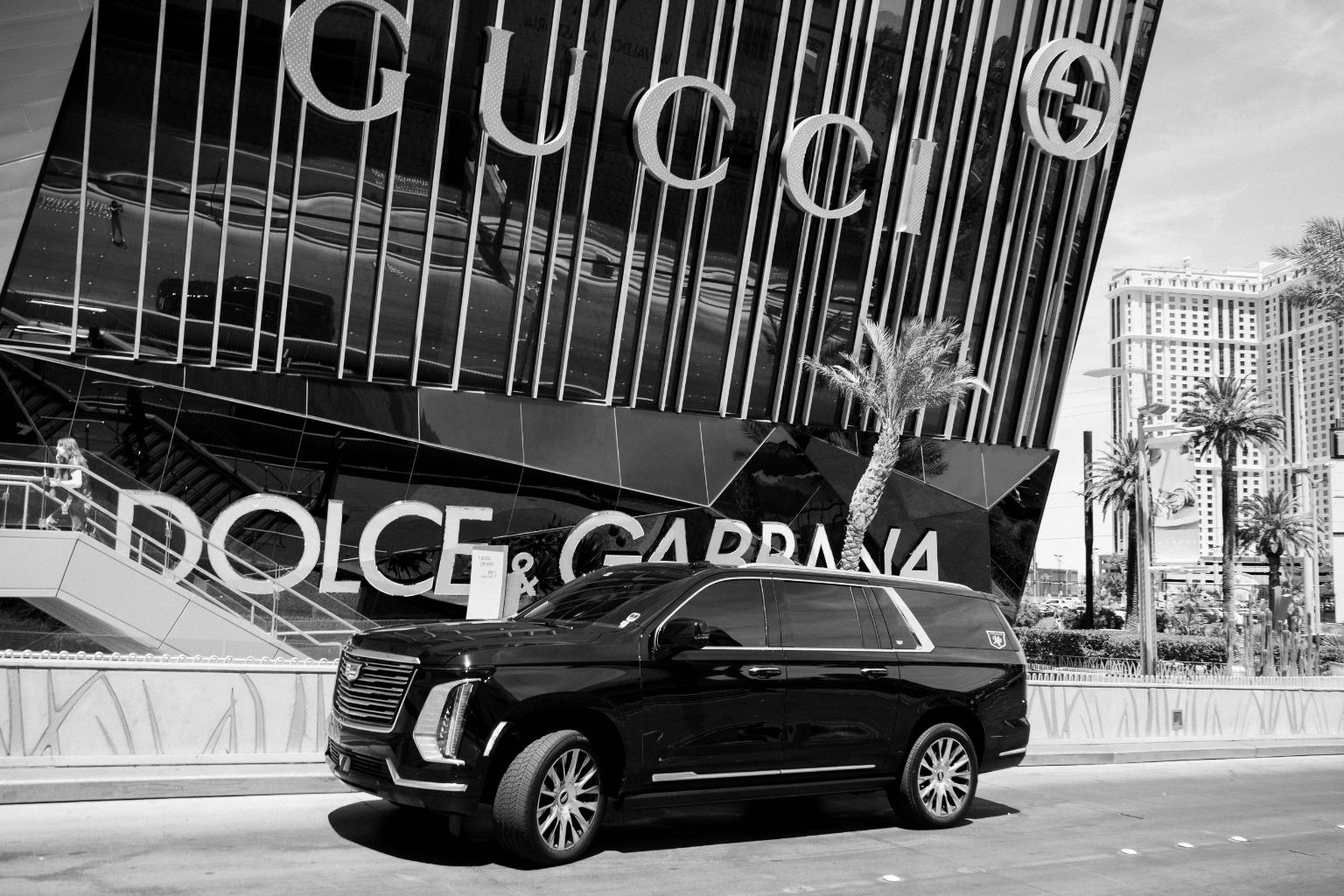 Black-and-white photo of a black Cadillac Escalade with MP Limousine branding parked in front of a luxury retail building displaying Gucci and Dolce & Gabbana storefront signage in Las Vegas.