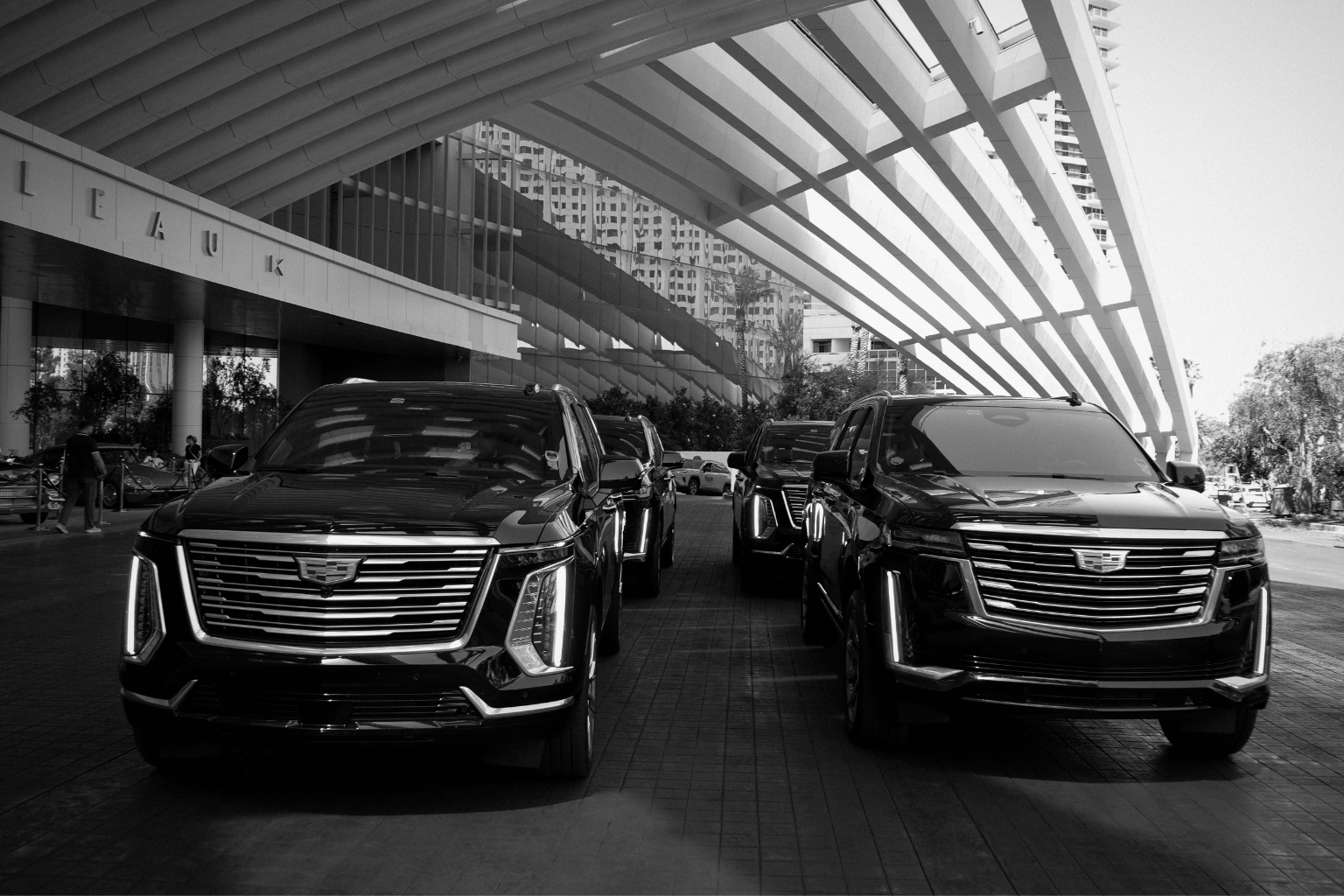 Black-and-white photo of two black Cadillac Escalades parked front-facing under the angular porte-cochère of the Fontainebleau, with additional fleet vehicles behind them.