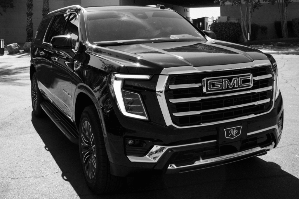 Black-and-white three-quarter front view of a black GMC Yukon with an MP Limousine branded front license plate, parked outdoors with the chrome GMC grille prominently featured.