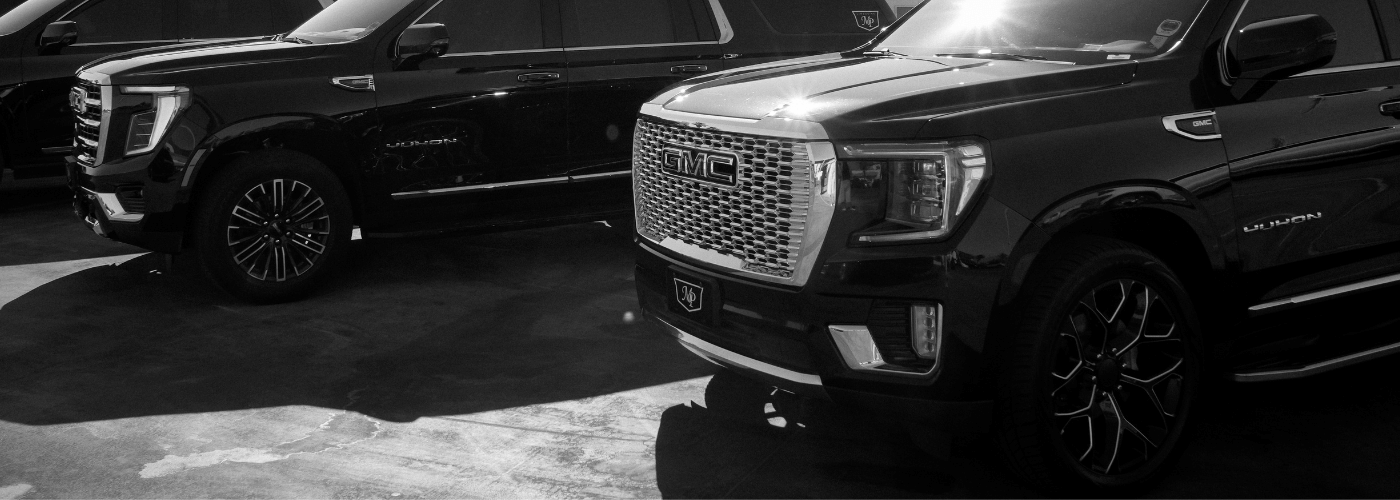 Black-and-white photo of two black GMC Yukon SUVs parked side by side, with the chrome GMC grille and MP Limousine logo badge prominently featured on the front vehicle.
