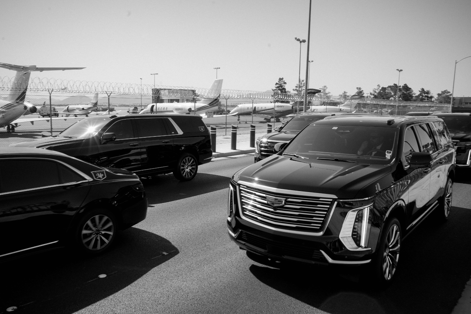 Black-and-white photo of multiple black Cadillac Escalades and SUVs from the MP Limousine fleet staged on a roadway beside a private jet terminal, with parked aircraft visible behind the perimeter fence.