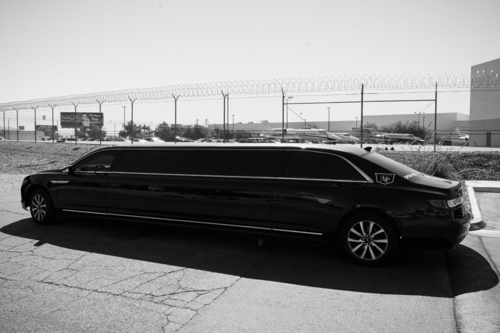 Black-and-white side profile of a black stretch limousine with MP Limousine branding on the rear door, parked outdoors near an airfield with private jets visible in the background.