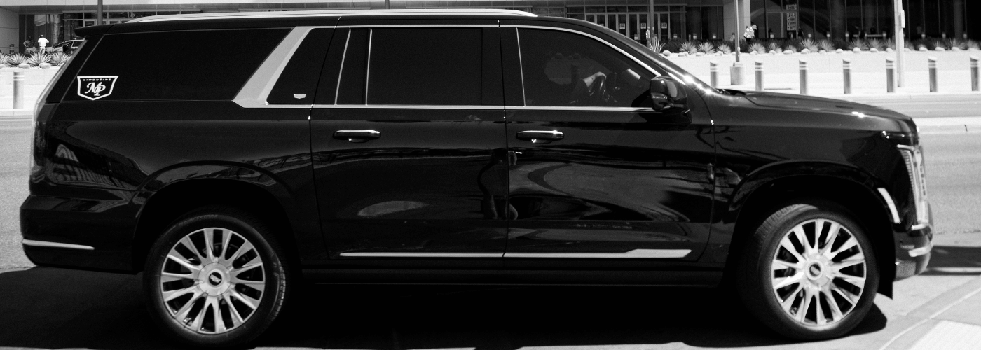 Black-and-white side profile of a black Cadillac Escalade with the MP Limousine logo on the rear quarter panel, parked on a city street.